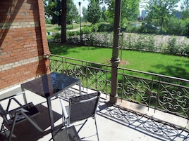Un balcón con mesa y sillas de vidrio, una pared de ladrillo y vistas a un parque verde con barandilla de metal.