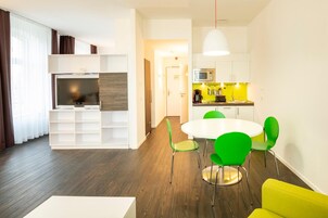 Una sala de estar moderna con un televisor de pantalla plana, una zona de comedor con una mesa blanca y sillas verdes, y una cocina con gabinetes blancos y salpicadero amarillo.