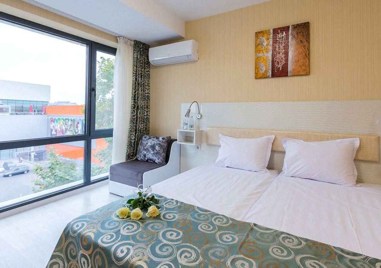 A hotel room with a large bed, a bedside table, a chair, a window with curtains, and a wall-mounted air conditioner.