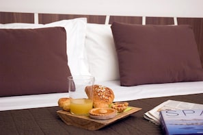 A breakfast setup on a wooden tray with pastries, a glass of orange juice, and a magazine on a bed with white and brown pillows.