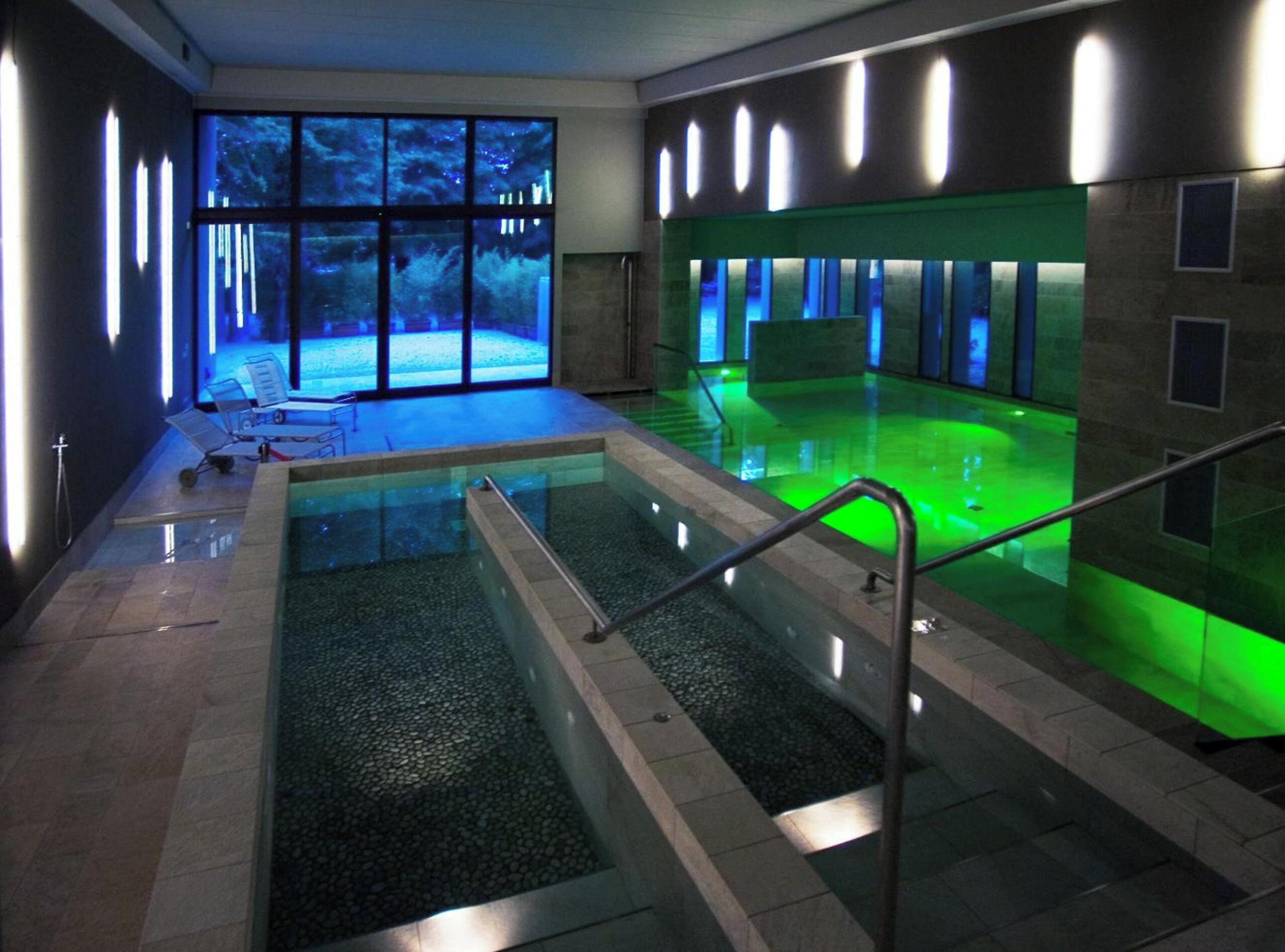 Une piscine intérieure avec une paroi en verre, un espace détente avec des fauteuils et une vue sur une forêt grâce à de grandes fenêtres.