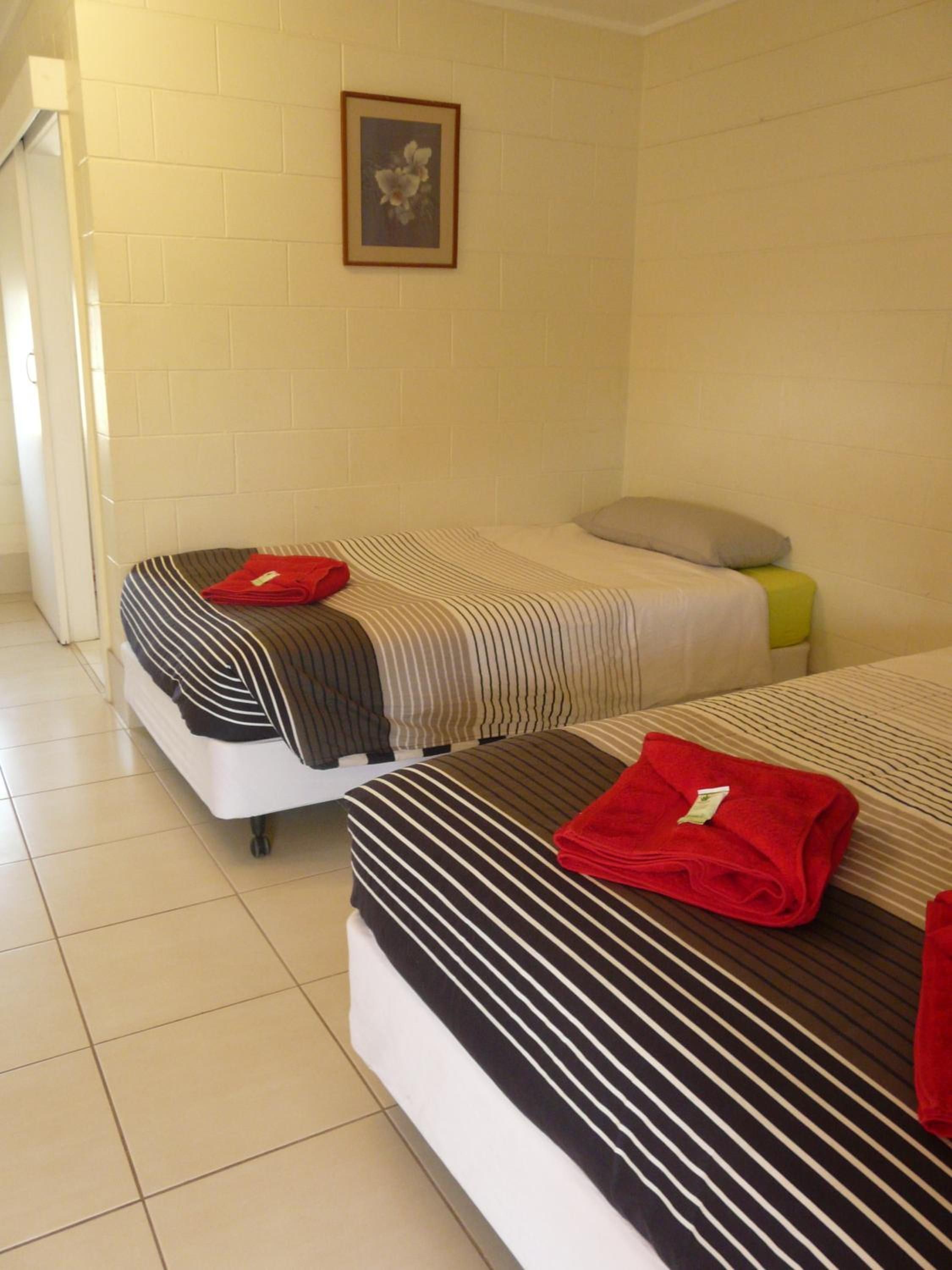 Quarto de hotel com duas camas, cada uma com roupa de cama listrada e uma toalha vermelha. Há um quadro na parede e um travesseiro amarelo em uma das camas.