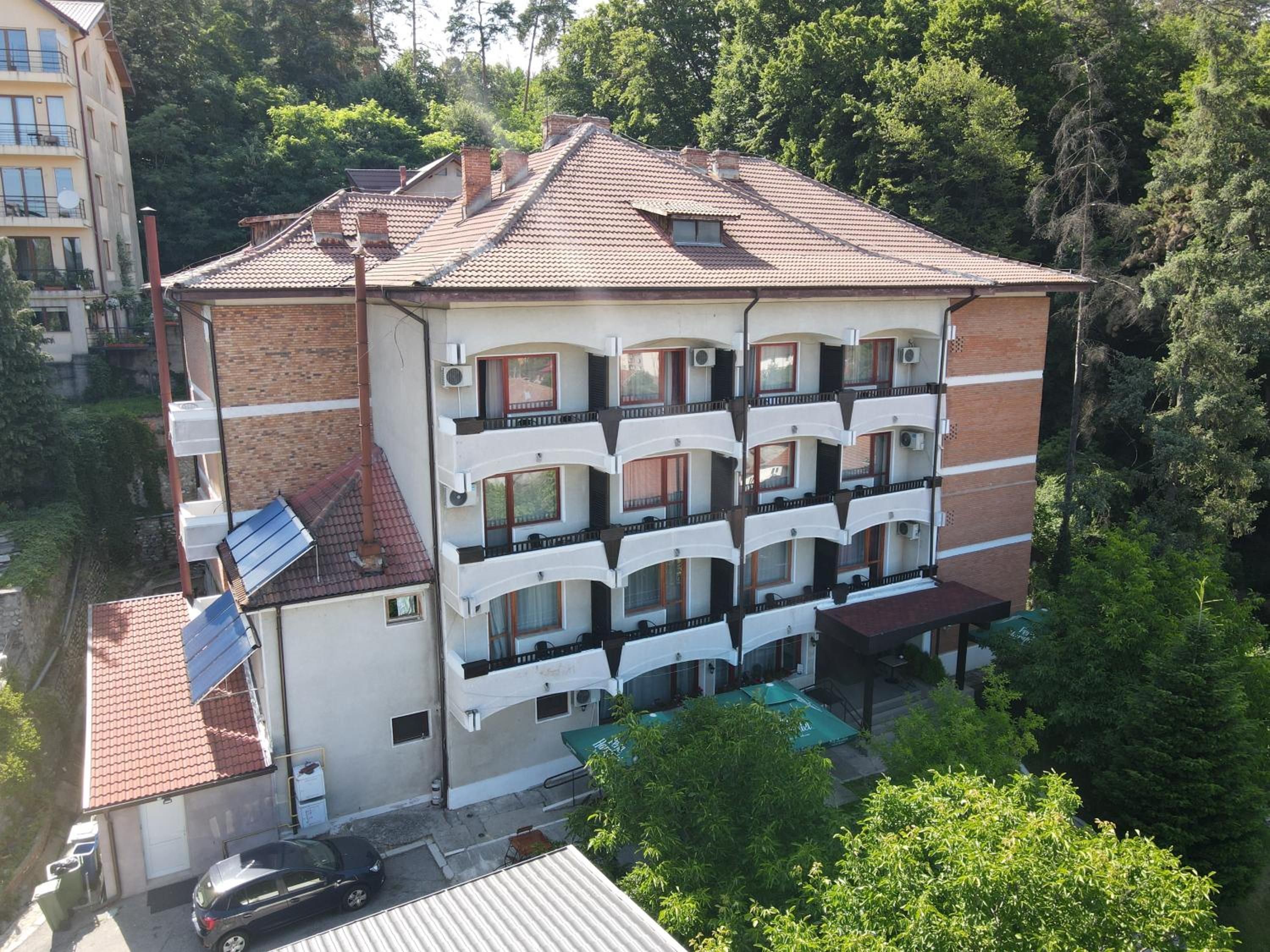 A multi-story building with balconies, a car parked in front, and greenery around.