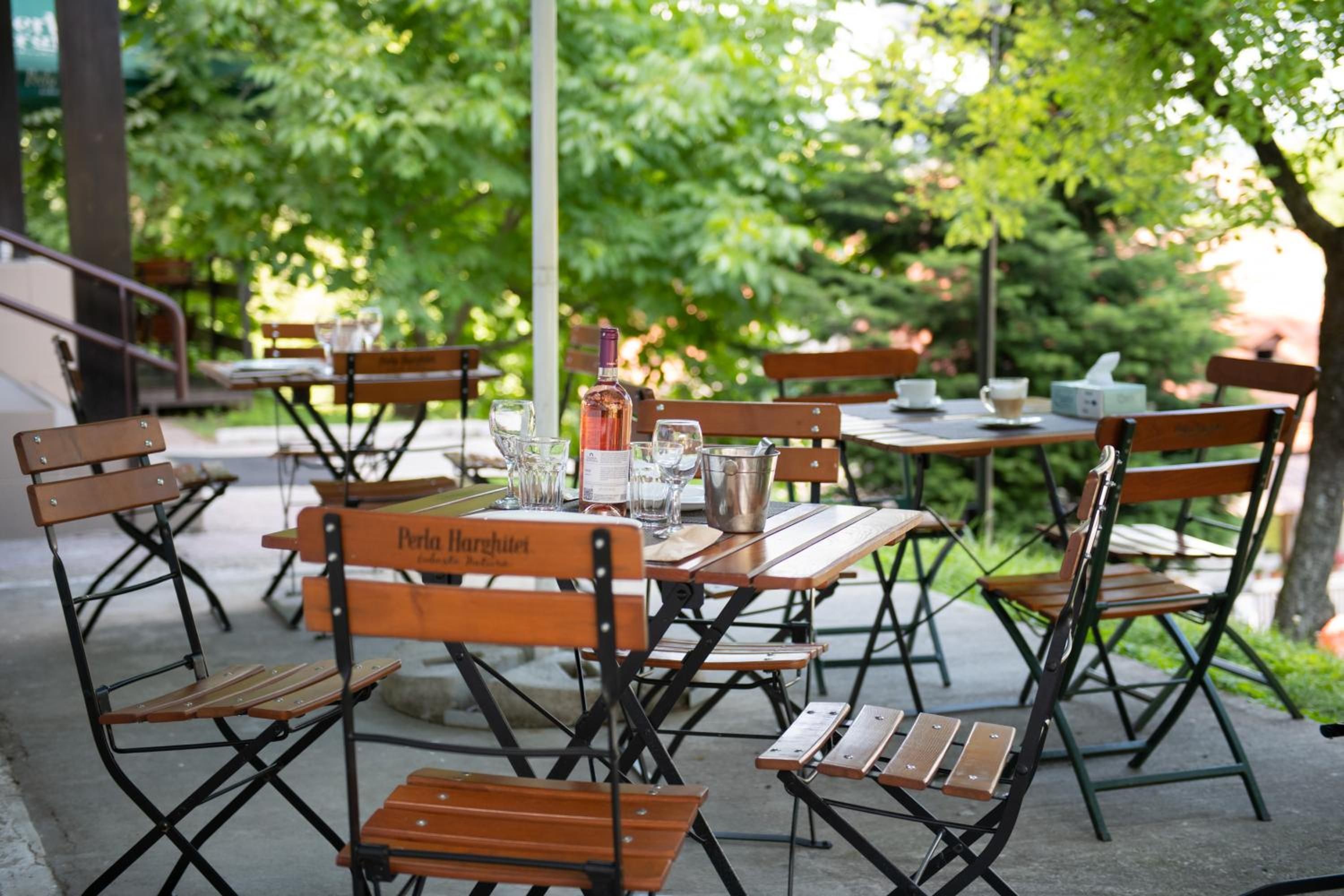 Outdoor tables and chairs with a bottle and glasses, surrounded by greenery.