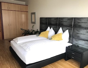A bedroom with a bed, a nightstand, a wooden wardrobe, and a plant. - Hotel Bellini (Leoben)