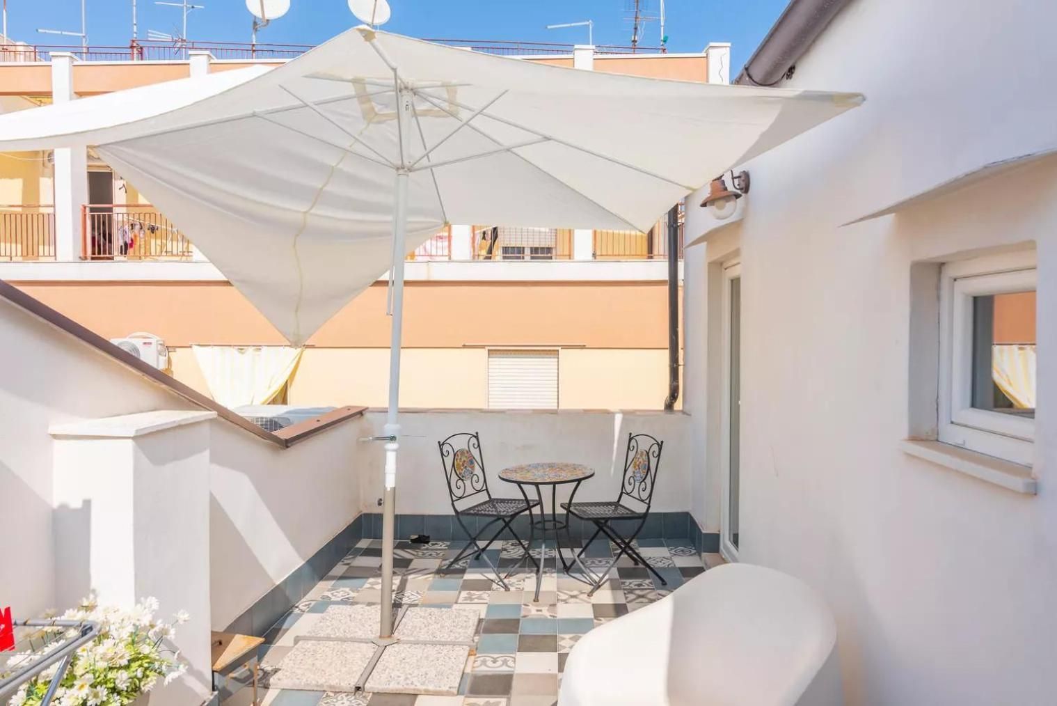 Un piccolo patio esterno con tavolo e due sedie, un ombrellone bianco e un pavimento piastrellato.