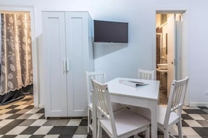 Una habitación con una mesa y sillas blancas, un televisor montado en la pared y un armario.