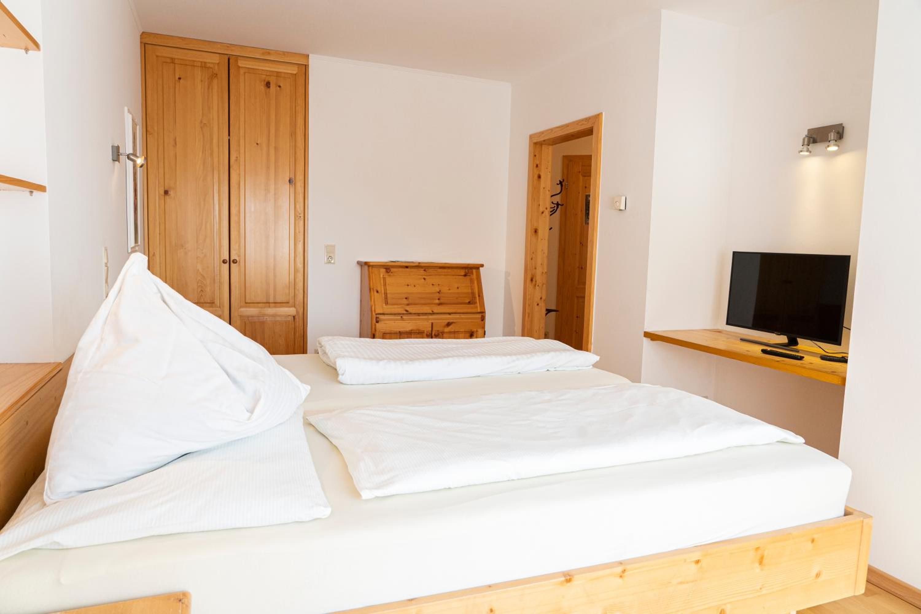 A bedroom with a bed, a wooden dresser, a television on a shelf, and a wardrobe.