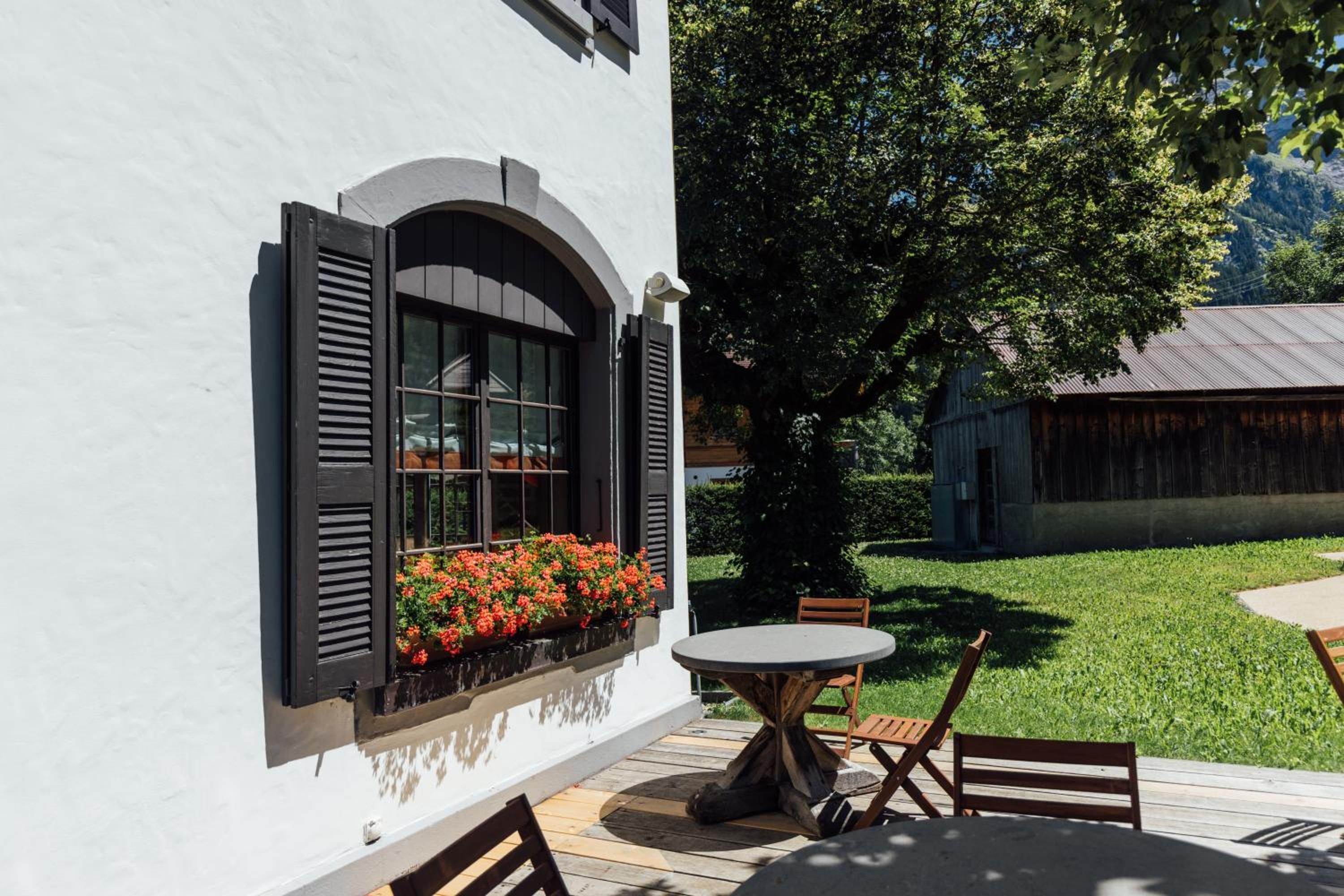 Ein weißes Gebäude mit einem Fenster, das mit schwarzen Rollläden und einem Blumenkasten mit roten Blumen versehen ist.