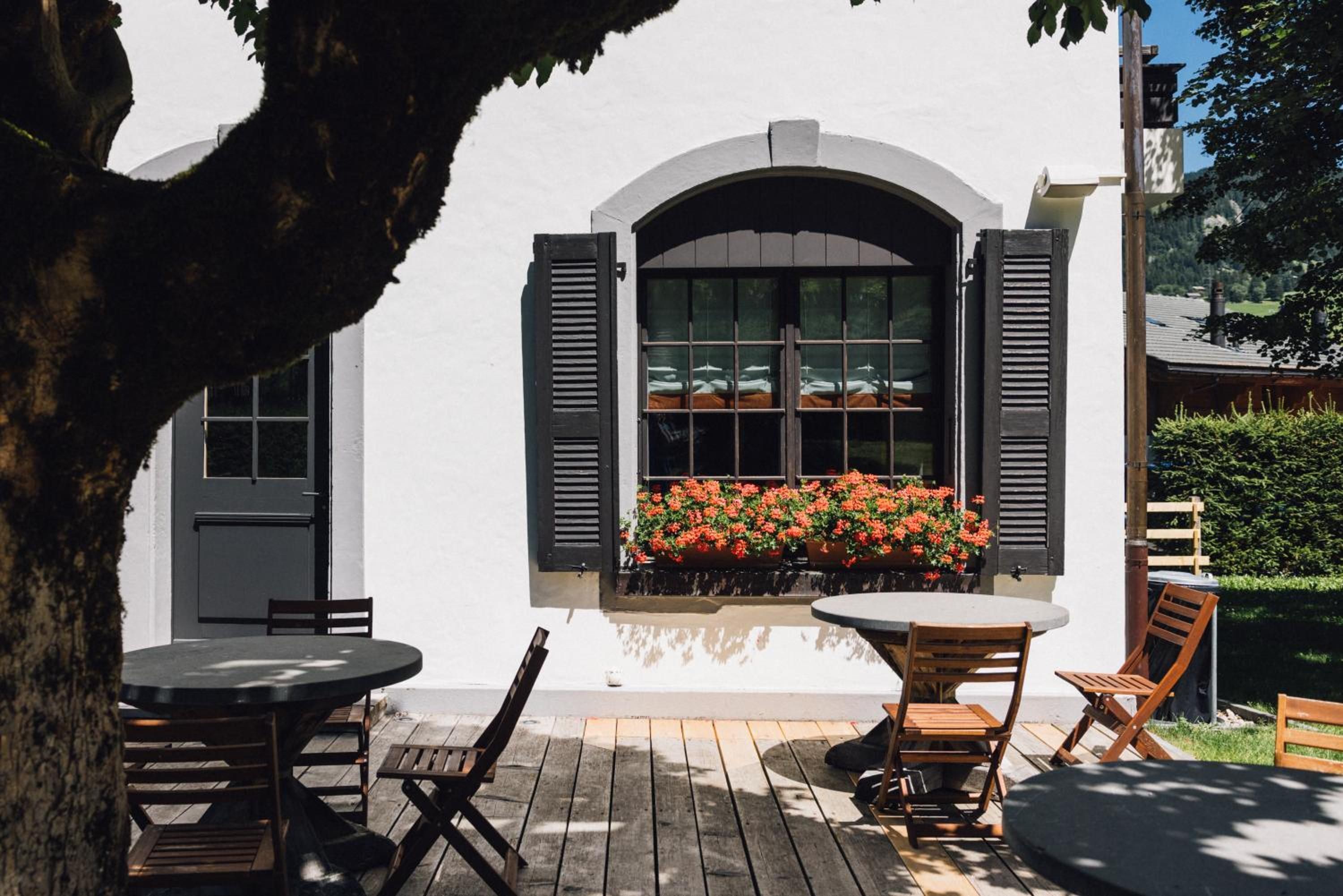 An outdoor seating area with tables and chairs, a window with shutters, and a view of a garden.