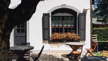 Ein Sitzbereich im Freien mit Tischen und Stühlen, ein Fenster mit Rollläden und Blick auf einen Garten.