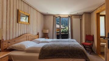 Ein Schlafzimmer mit einem hölzernen Bett, einem Nachttisch, einem Sessel und Blick auf eine gebirgige Landschaft durch ein großes Fenster.