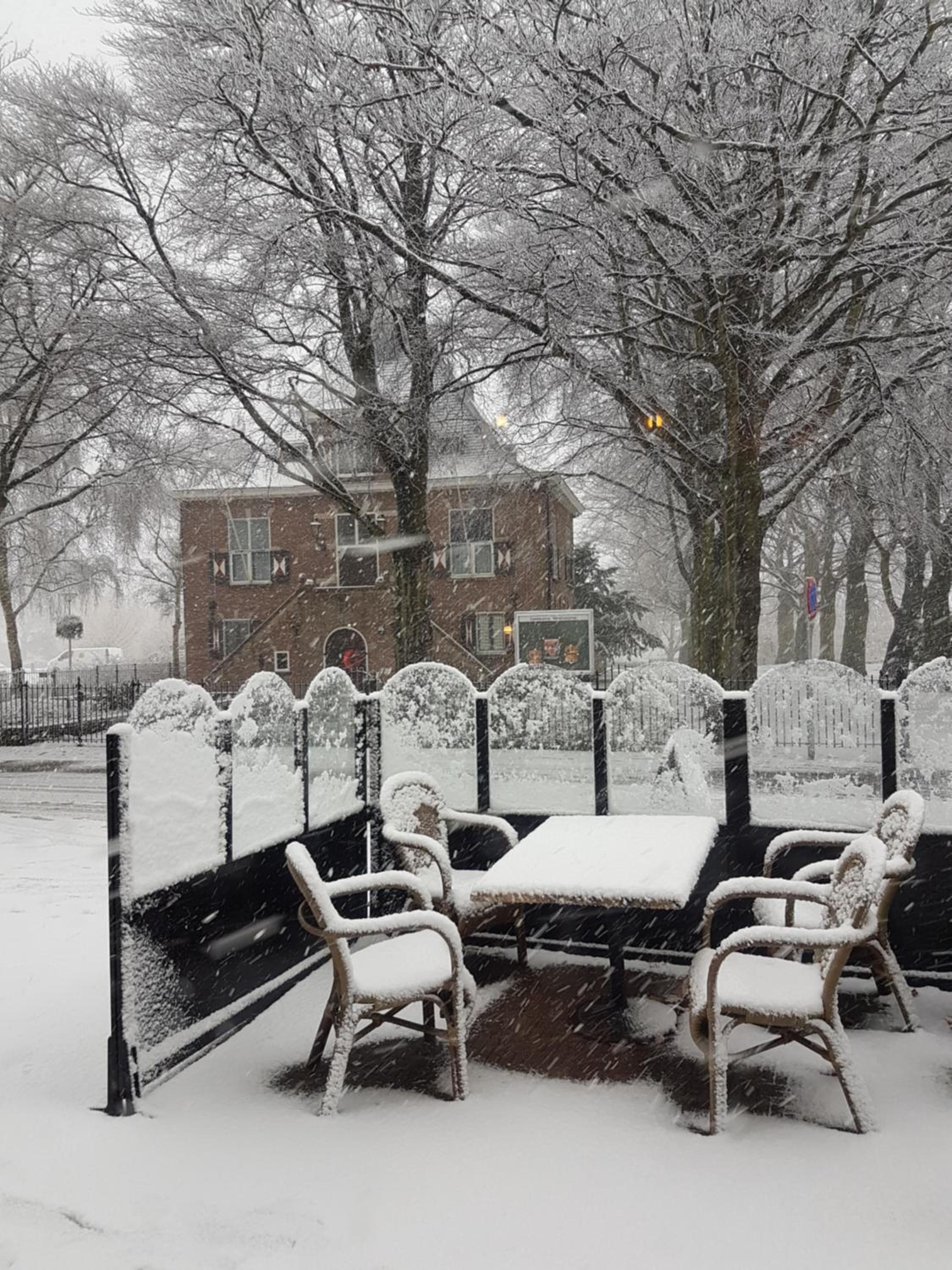 A snow-covered outdoor seating area with a table and chairs, surrounded by trees and a building in the background.