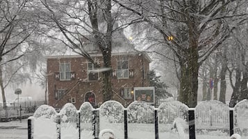 A snow-covered outdoor seating area with a table and chairs, surrounded by trees and a building in the background.