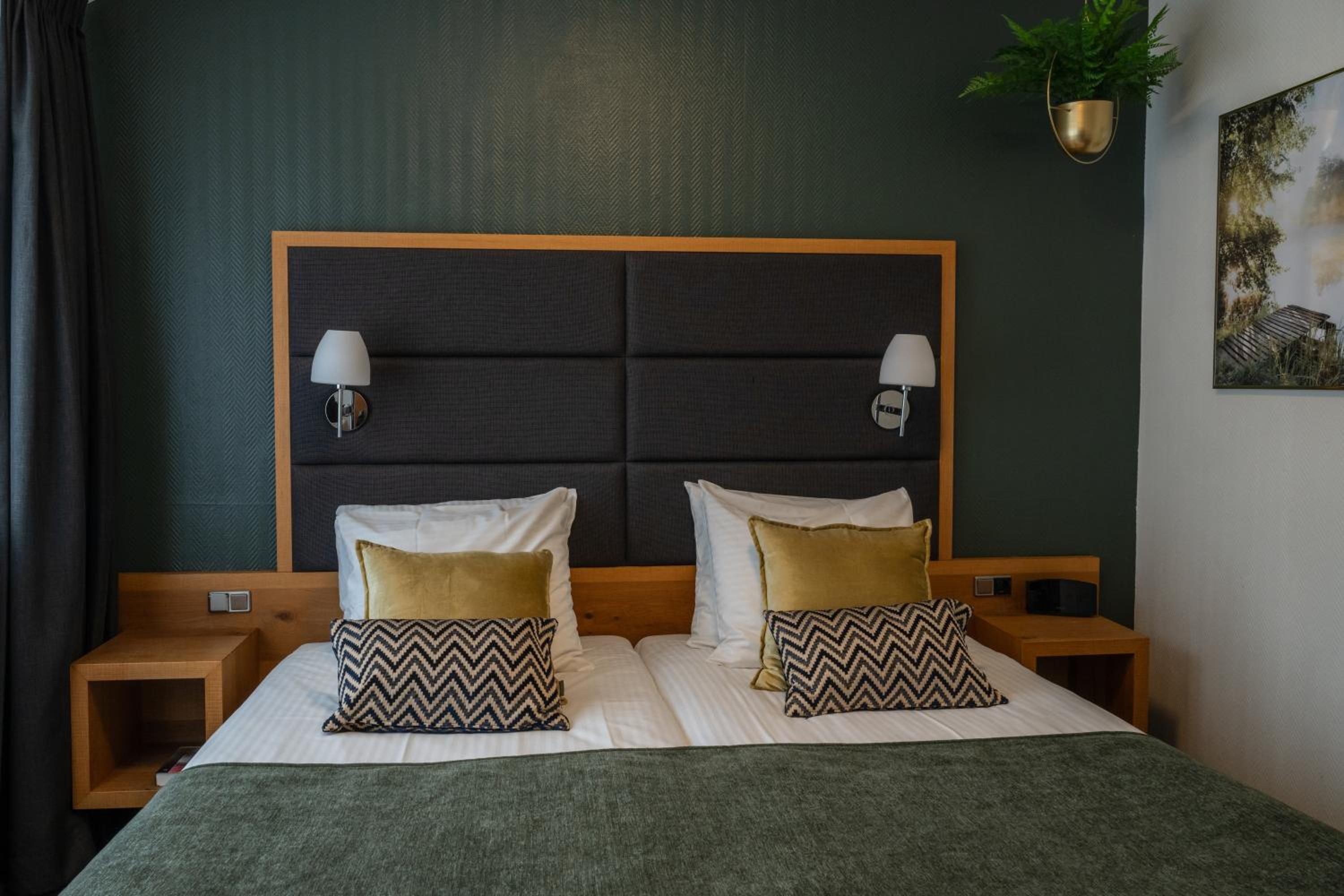 Una cama doble con dos mesitas de noche, un cabecero con lámparas de pared y una planta verde en la pared.