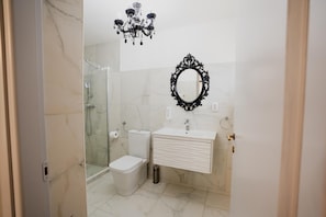 A bathroom with a glass shower enclosure, a toilet, a sink with a mirror, and a chandelier.