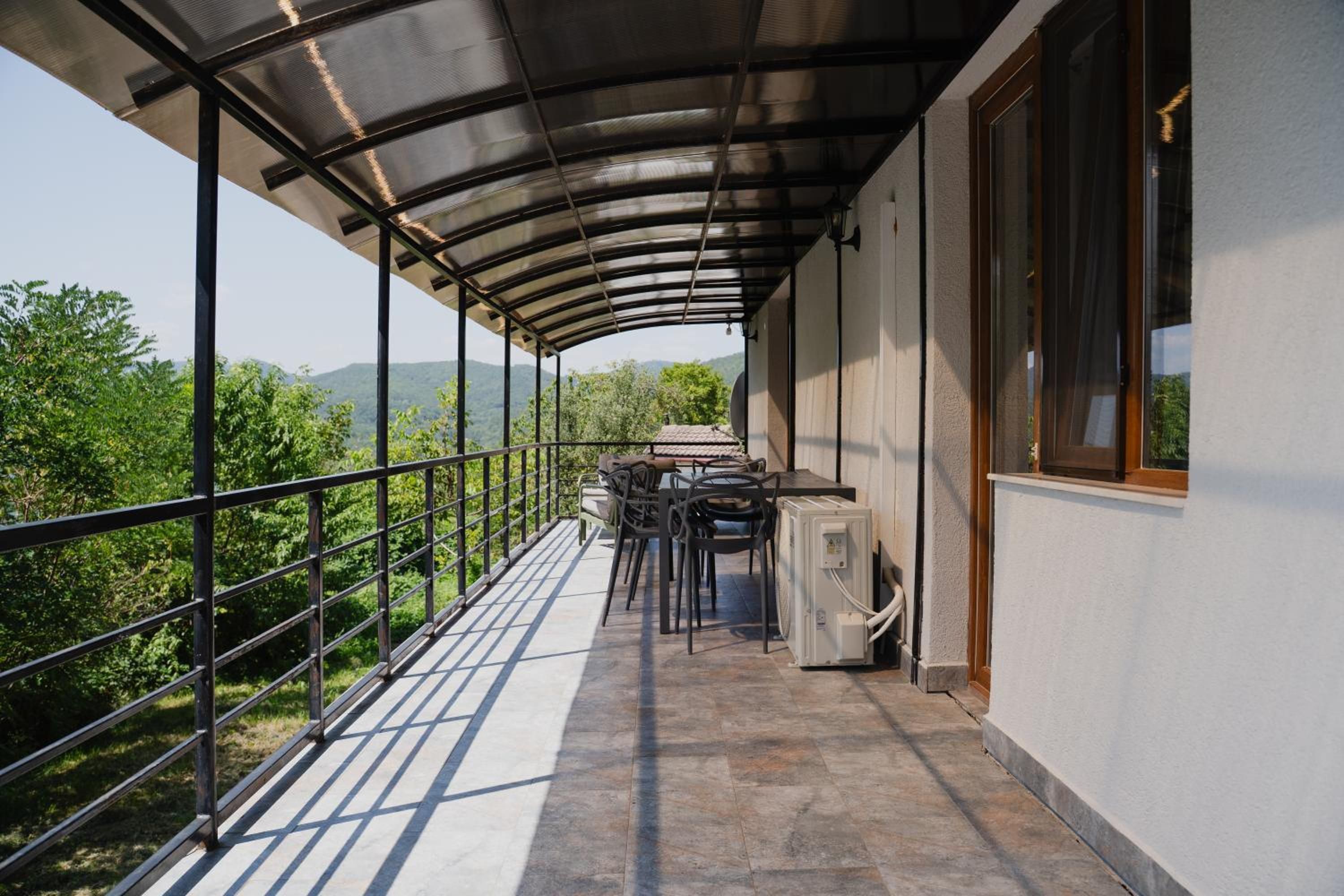 A covered outdoor balcony with a table and chairs, a window, and a view of greenery and hills.