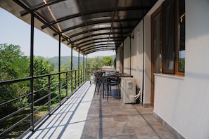 A covered outdoor balcony with a table and chairs, a window, and a view of greenery and hills.