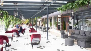 An outdoor cafe with tables and chairs, a covered seating area, and greenery.