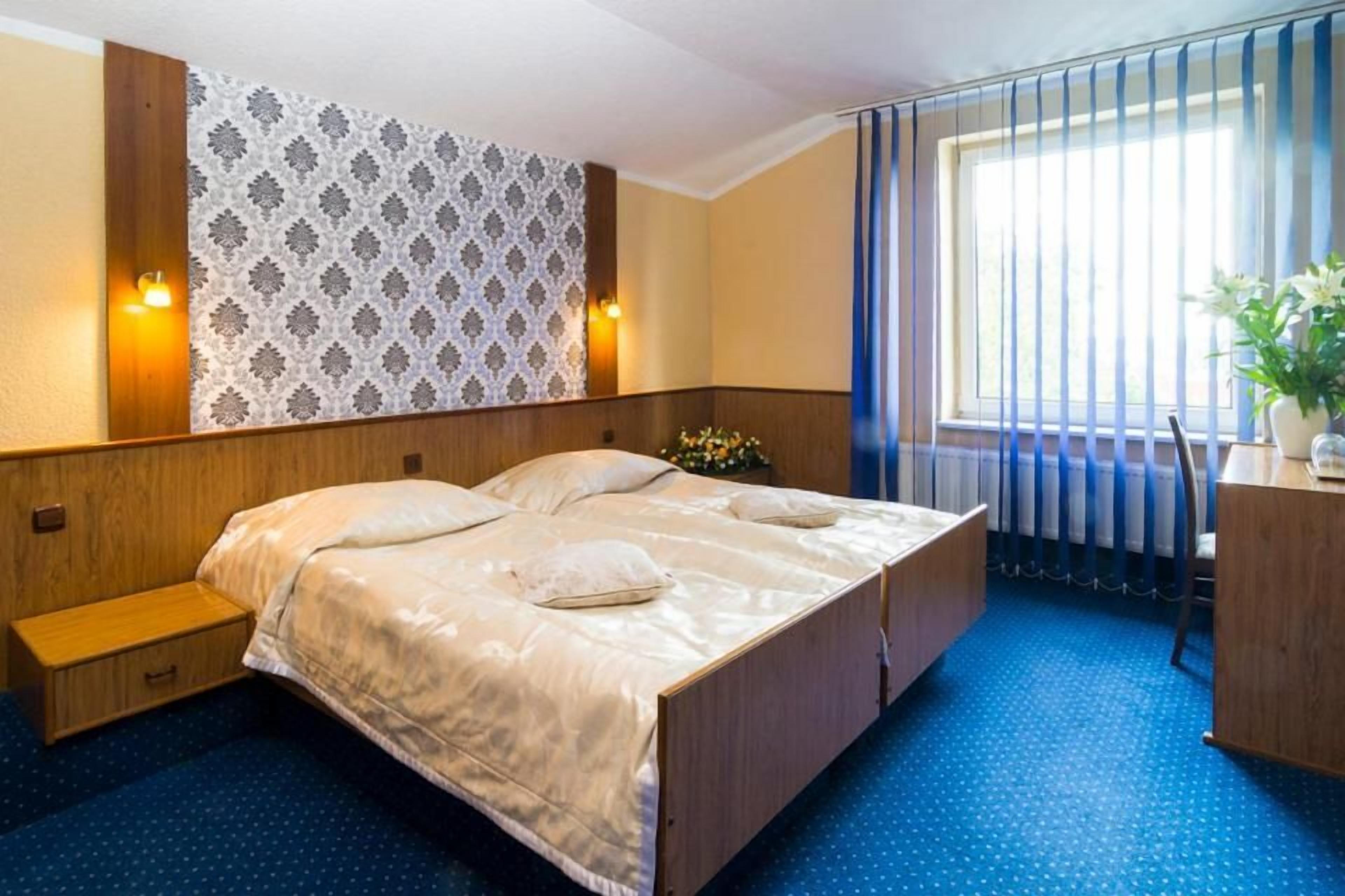 Ein Hotelzimmer mit einem Doppelbett, einem Holzkopfstück, einem kleinen Nachttisch, einer Ablagefläche, einem Fenster mit Vorhängen und einer Wand mit gemustertem Tapetenpapier.