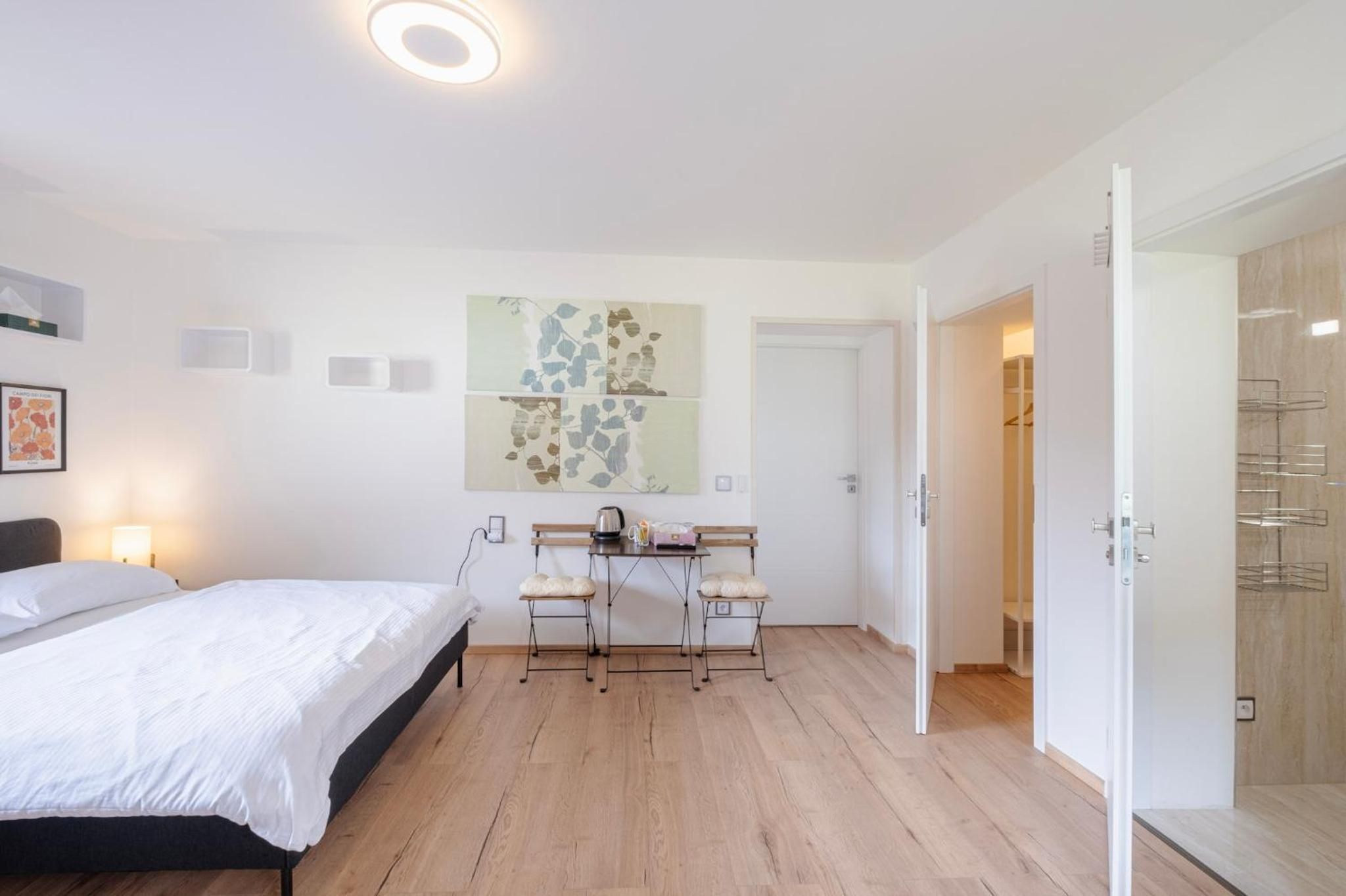 A bedroom with a bed, a desk with chairs, a coffee maker, and a wardrobe.