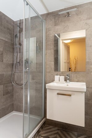 Un baño moderno con ducha de vidrio, un lavamanos blanco con espejo encima y baldosas de piso estampadas.