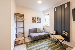 Una habitación con sofá, una silla, una estantería de madera con ropa, una mesita y dos láminas botánicas enmarcadas en la pared.