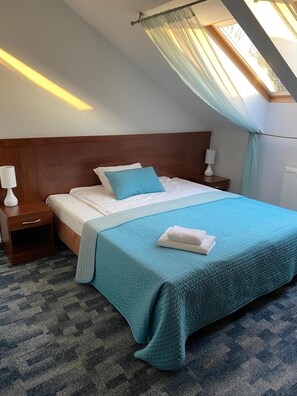 Quarto de hotel com cama, mesa de cabeceira, luminária, janela com cortina e radiador.