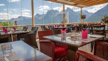 An outdoor restaurant with tables and chairs, large windows offering mountain views, and a wooden ceiling structure.