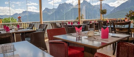 An outdoor restaurant with tables and chairs, large windows offering mountain views, and a wooden ceiling structure.