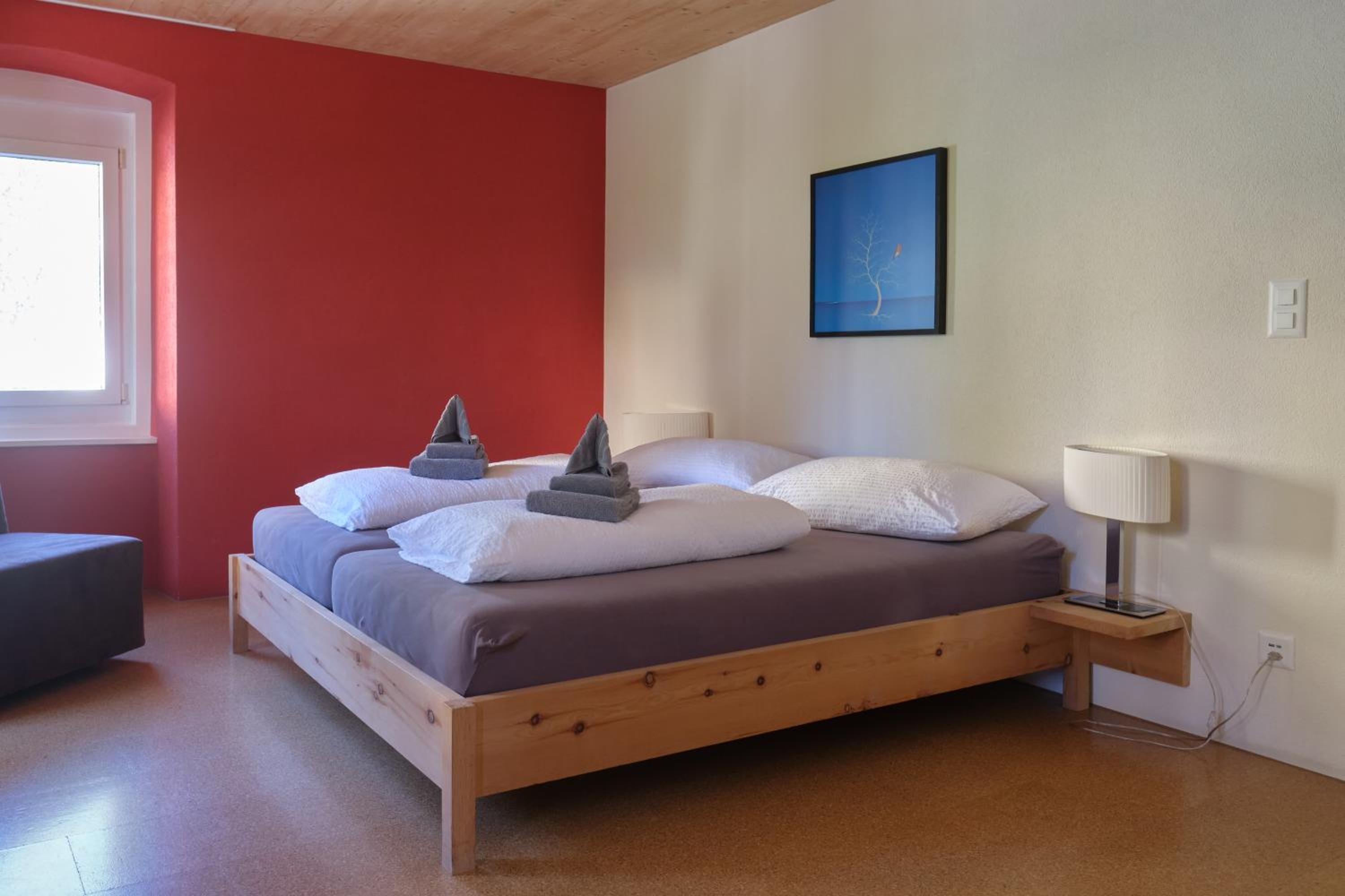 A bedroom with a wooden bed, a bedside table with a lamp, a window, and a wall with a framed picture.