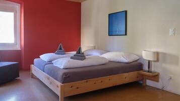 A bedroom with a wooden bed, a bedside table with a lamp, a window, and a wall with a framed picture.
