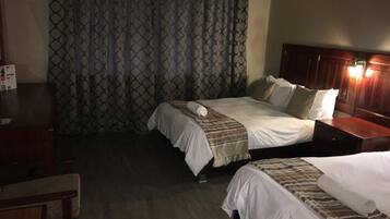 Une chambre d’hôtel avec deux lits, une tête de lit en bois, un bureau et un rideau orné d’un motif géométrique.