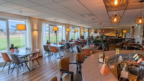 Un restaurante con varias mesas y sillas, una zona de bar y grandes ventanales con vistas al exterior.