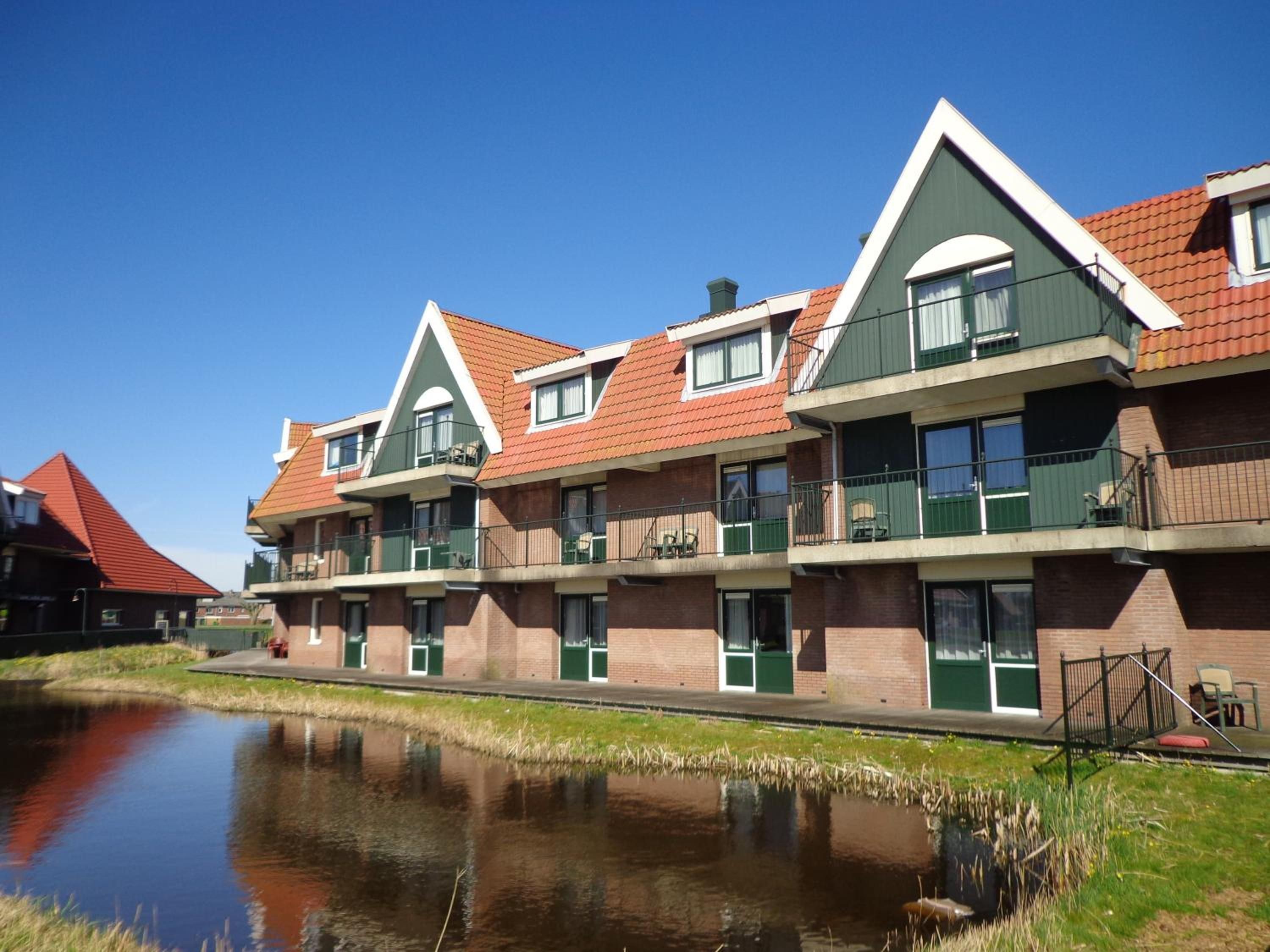 En række moderne bygninger med flere etager og altaner, en kanal foran og en klar, blå himmel.