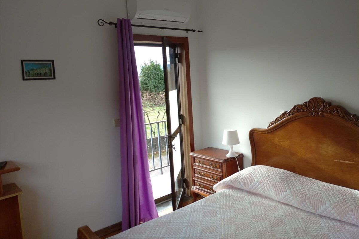 A bedroom with a bed, a dresser, a nightstand, a window with purple curtains, and an air conditioning unit.