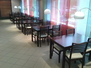 Comedor con varias mesas y sillas, amplias ventanas con cortinas e iluminación colgante.