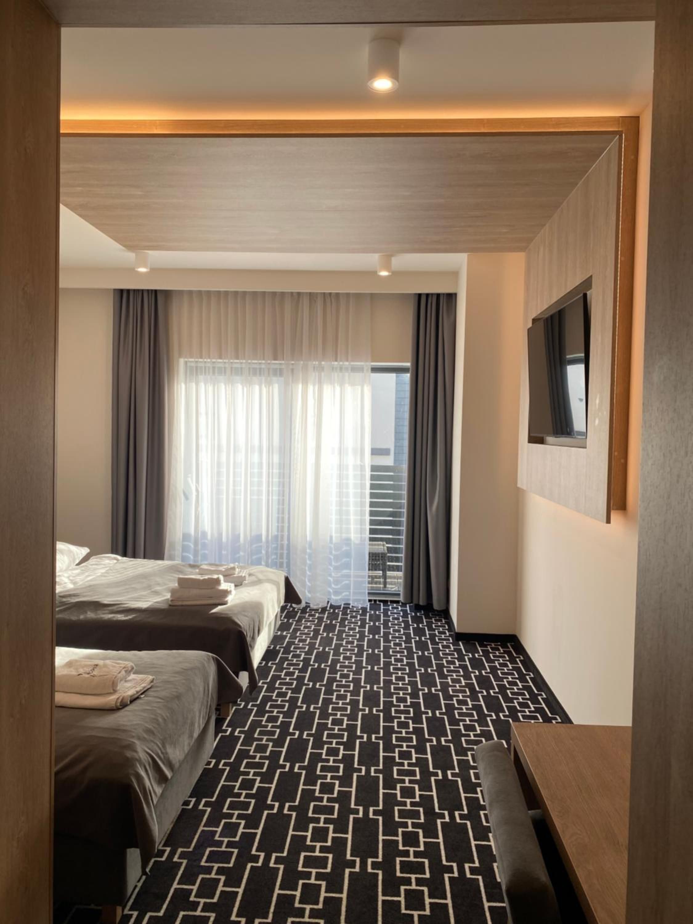 A hotel room with two beds, a patterned carpet, a flat-screen TV, and a window with curtains.