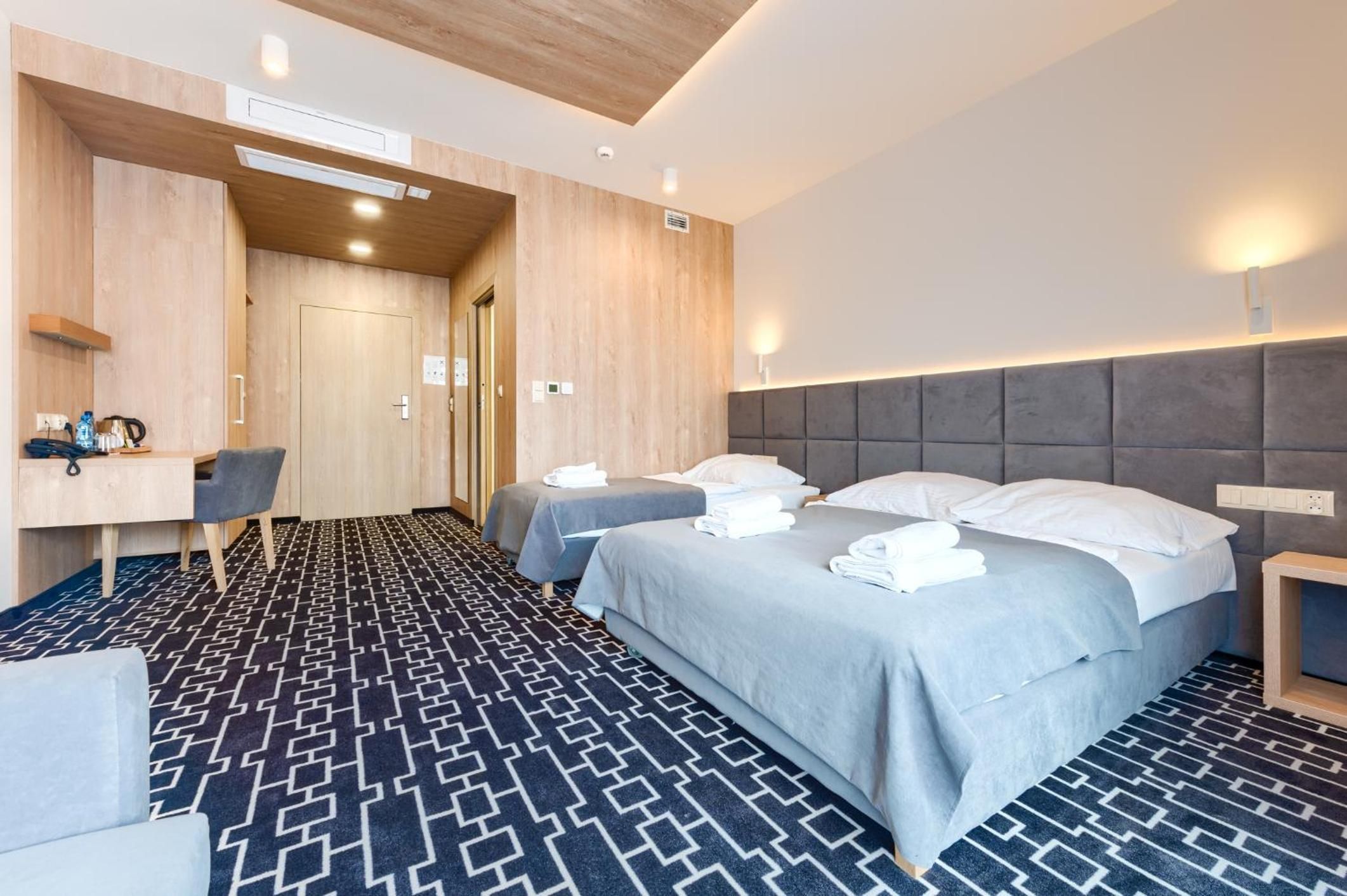 A hotel room with a bed, two bedside tables, a desk with a chair, and a patterned carpet.