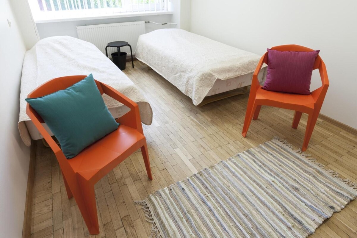 A small room with two single beds, a small round table, two orange chairs, and a striped rug.