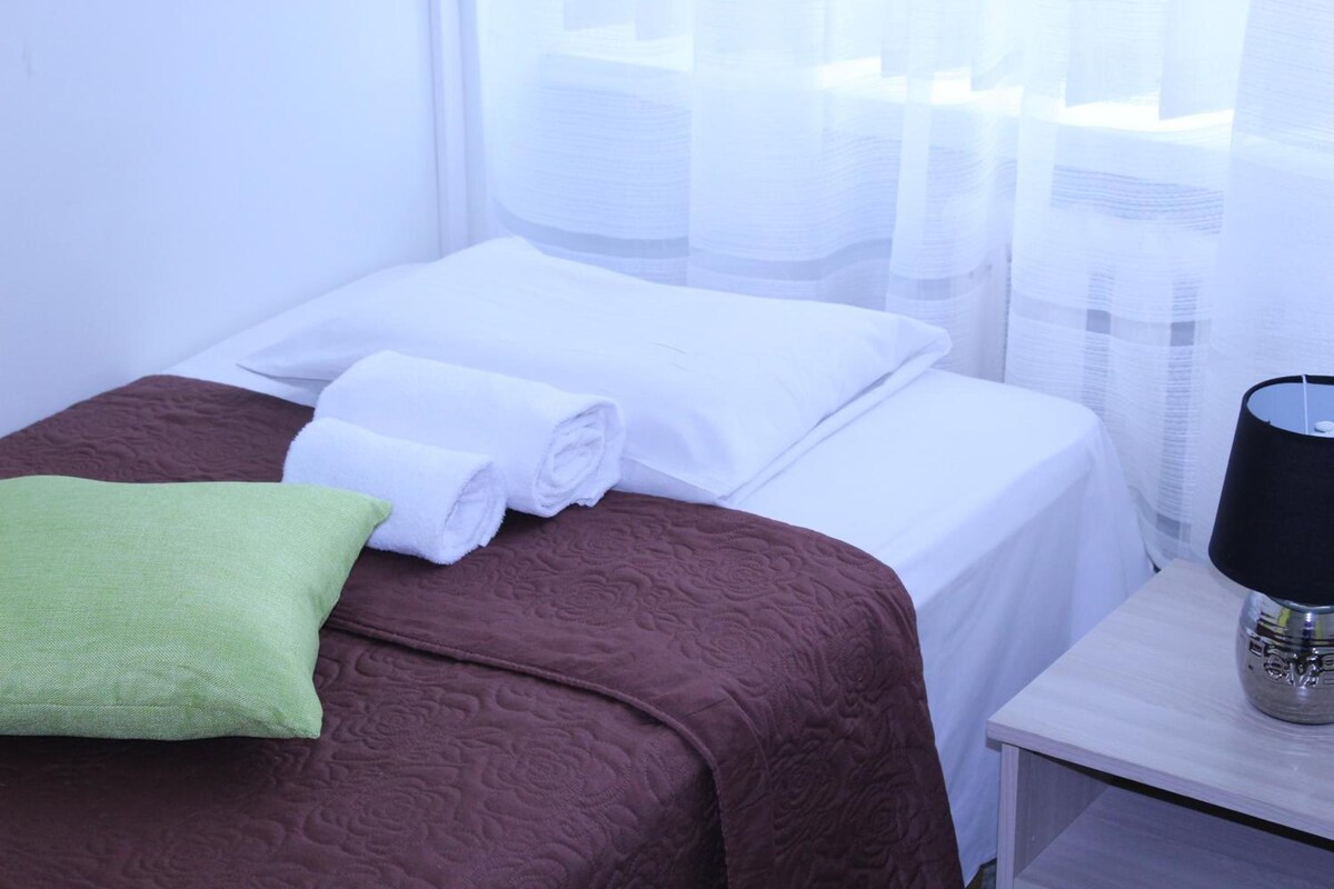 A hotel room with a neatly made bed, a green pillow, and a bedside lamp.