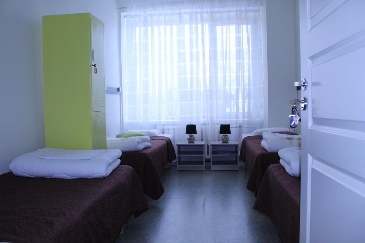 A hotel room with two beds, a wardrobe, a window with sheer curtains, bedside tables with lamps, and a door.