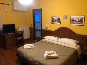 Habitación de hotel con una cama, un escritorio con televisor, una silla, un radiador y dos cuadros en la pared.