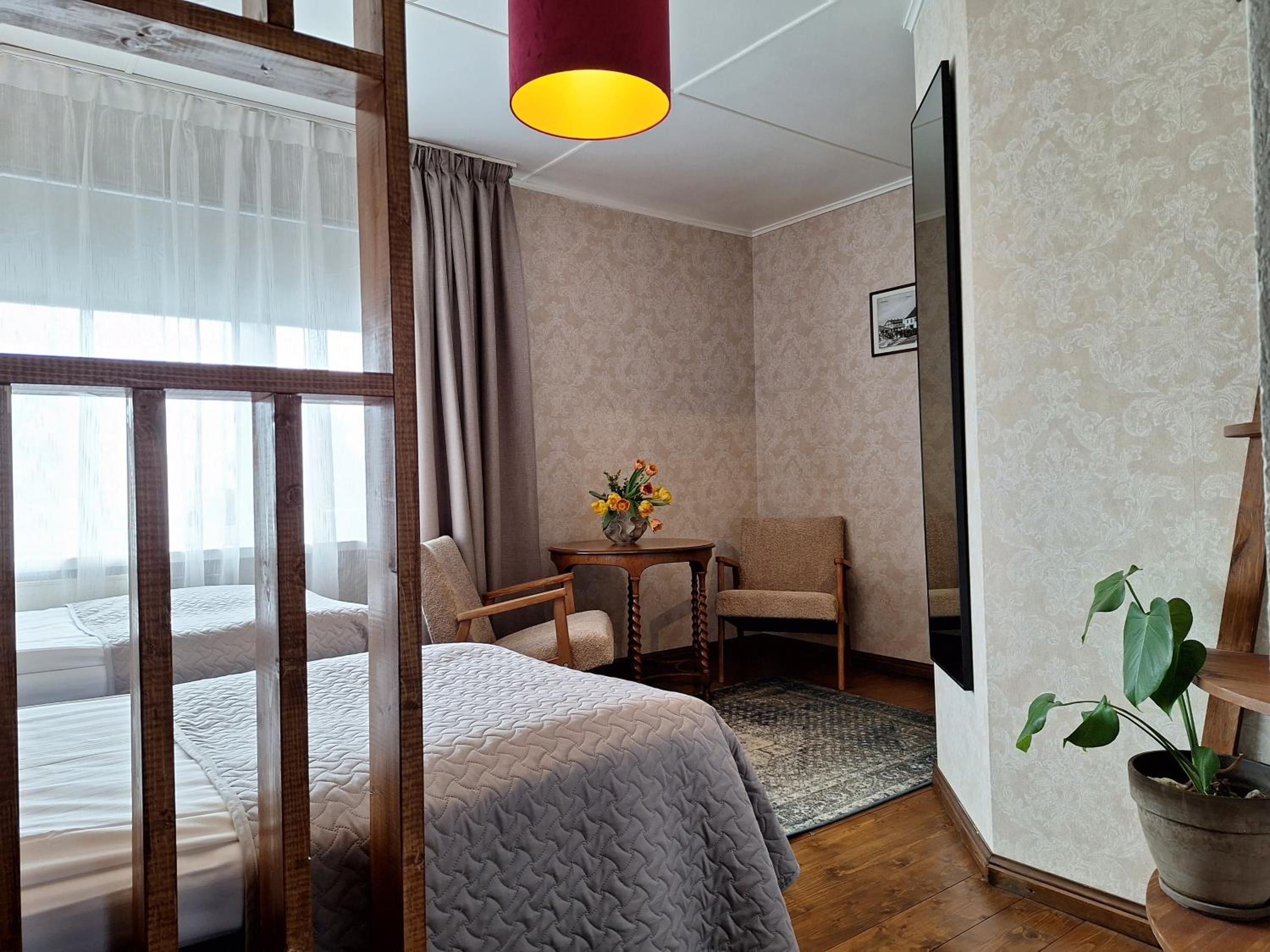 A hotel room with a bed, two chairs, a small table with flowers, a potted plant, and a large mirror.