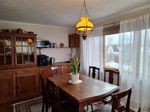 Un comedor con mesa de madera, sillas, lámpara colgante y vista al exterior a través de una ventana con cortinas traslúcidas.