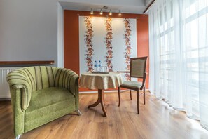 A room with a green upholstered armchair, a wooden chair, a small round table with a tablecloth, and a decorative wall with vertical floral patterns.