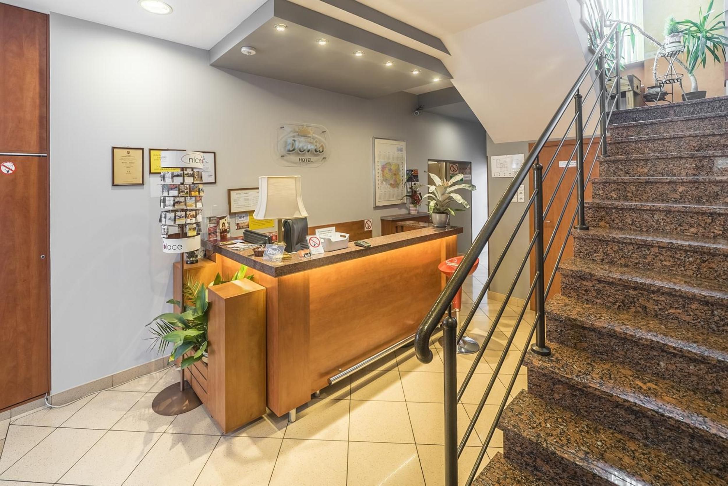 A hotel reception desk with a wooden counter, a computer, and a lamp. There is a staircase with a metal railing leading to an upper floor.
