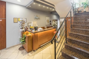 A hotel reception desk with a wooden counter, a computer, and a lamp. There is a staircase with a metal railing leading to an upper floor.
