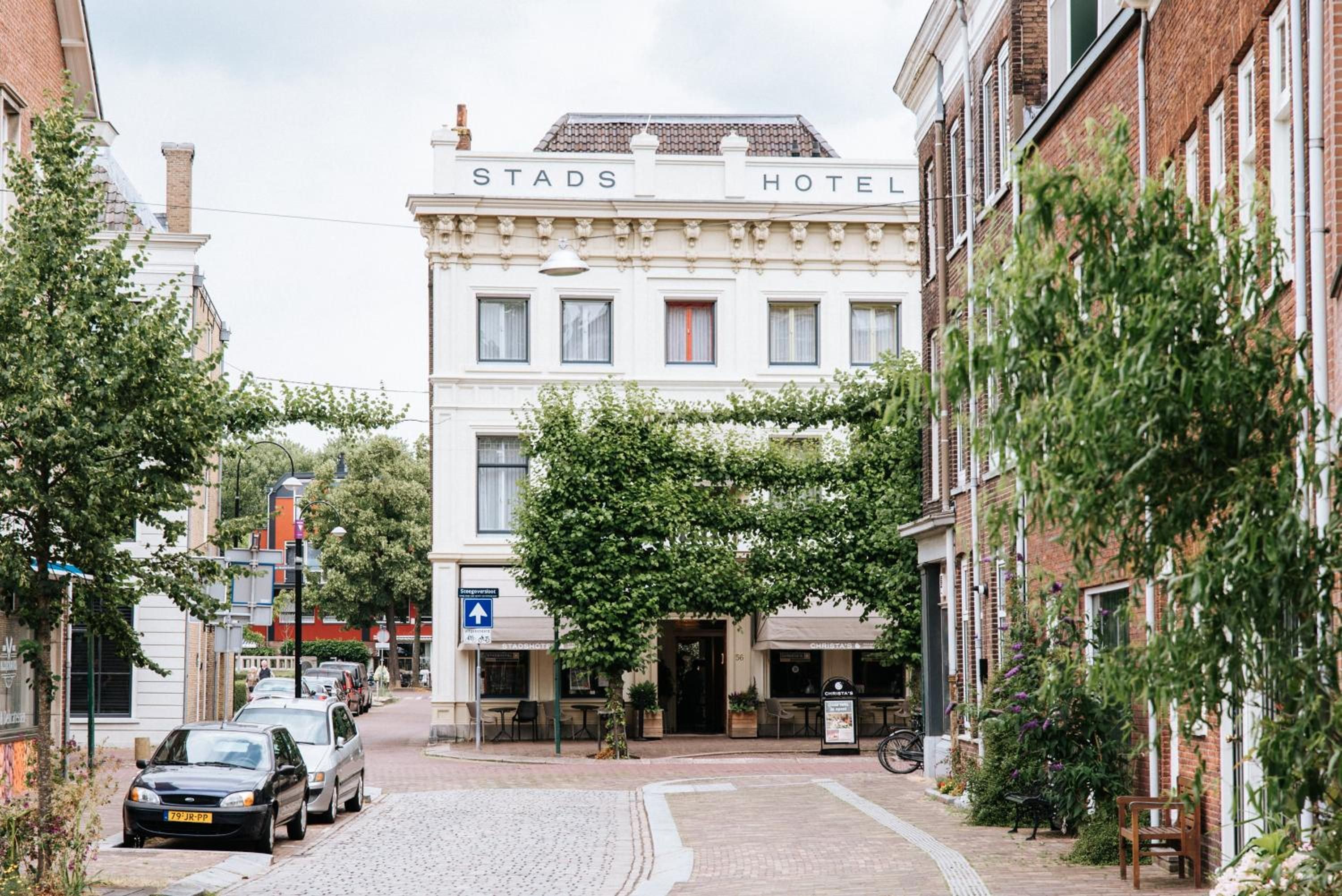 A street view of Stads Hotel with a cobblestone road, parked cars, and surrounding buildings.