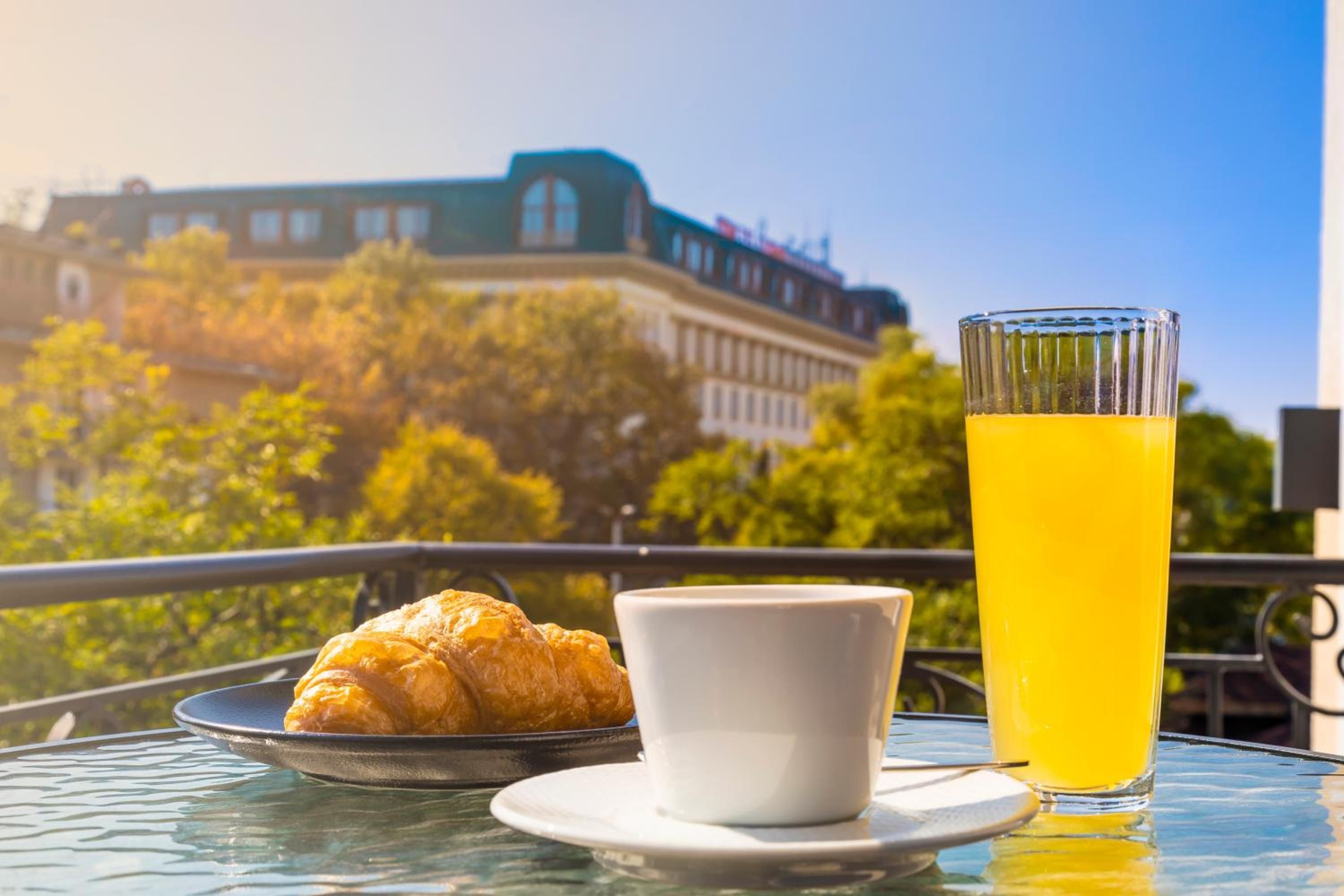 A table with a croissant, a cup, and a glass of orange juice, with a building and trees in the background.
